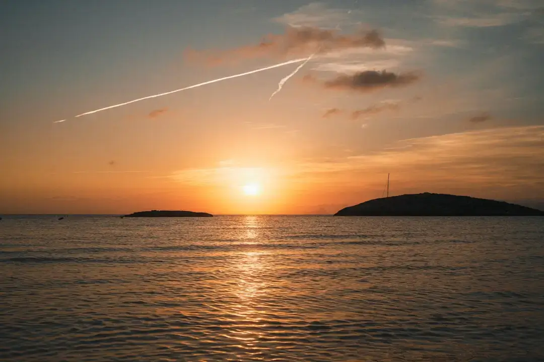 A vibrant orange and purple sunset over the crystal clear waters of an Ibiza beach with silhouette cliffs in the distance.