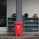 a red garbage can next to a shelf with objects on it