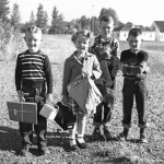 Four children with school bags and lunchboxes stand outside.