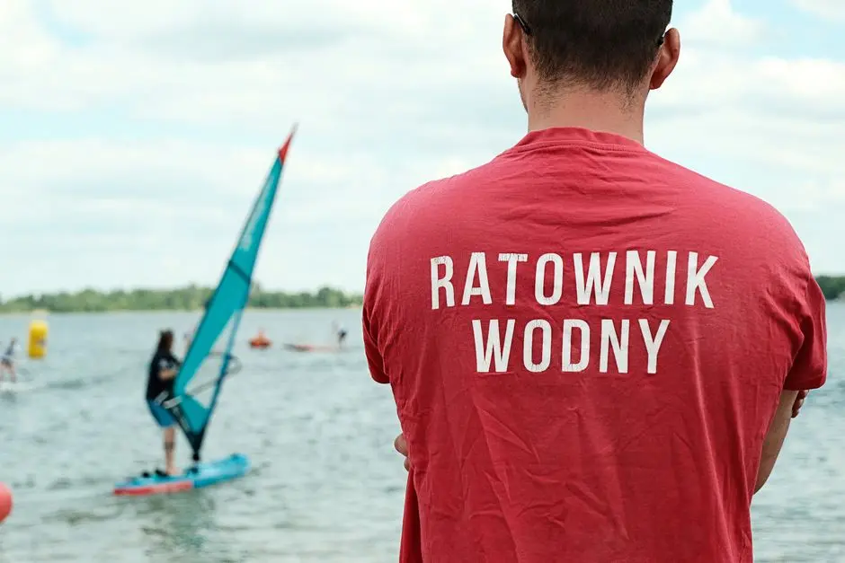 Ratownik medyczny nadzorujący pływaka na desce SUP na polskim jeziorze, pod częściowo pochmurnym niebem.